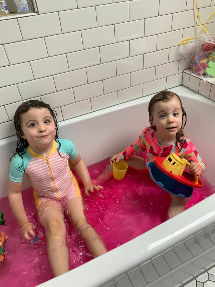 A young girl wearing a pink and aqua striped swimsuit sits in a bathtub filled with bright pink water. Her younger sister sits to her left in a pink and orange camouflage print bathing suit while holding a yellow cup and plastic yellow, orange, and black tugboat bath toy. 