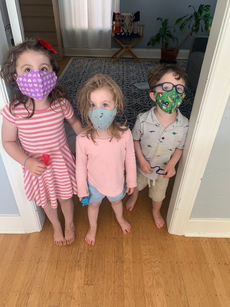A little girl standing on the left in a doorway wearing a purple cloth face mask peppered with hearts design and looking up. Her younger sister stands next to her left side, wearing a light blue cloth face mask, looks straight ahead. On the other side of the doorframe stands their young brother, wearing a green cloth face mask with a construction vehicle design, looking off to his left. 