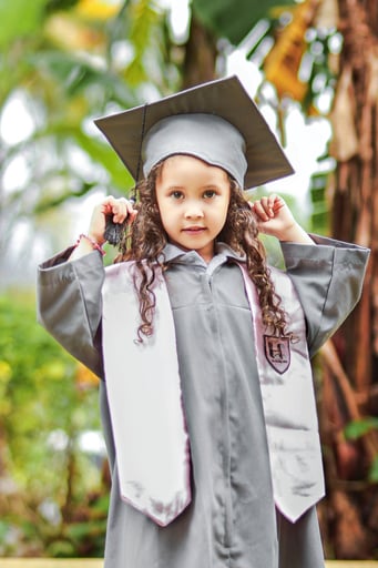 Preschool-aged girl standing in a silver-grey graduation gown and cap.