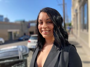 A smiling woman with long black hair wearing a black blazer, standing outdoors against a city backdrop with a blue sky.