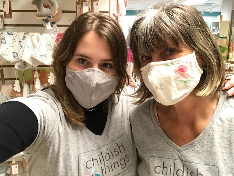 A mother is standing to the left of her teenage daughter. The mother is wearing a cream-colored mask with a pink roses pattern. The daughter is wearing a solid light grey mask and has her left arm draped on her mother’s shoulder, while her right arm is extended, holding out the camera to take this photo. Both women are wearing light grey, short-sleeved V-neck t-shirts with the Childish Things logo seen on the chest, leaning their heads in towards one another. 