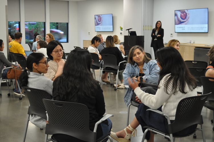 A speaker stands at the front of the room, while groups of parents sit and engage in conversation. 