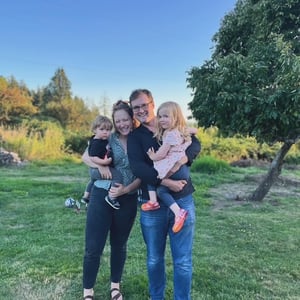 A man and woman standing in a grassy field on a sunny day, each holding a young child in their arms and smiling at the camera. 
