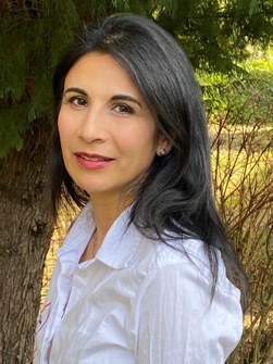 A portrait of a woman with long dark hair, wearing a white blouse, standing outdoors against a backdrop of trees.