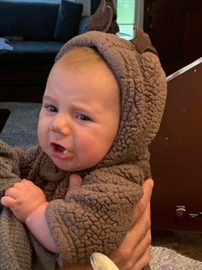 A baby wearing a hooded fuzzy brown outfit with a grouchy expression on their face.