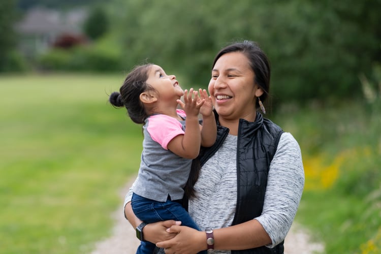 A Native American mom holds her toddler-age daughter affectionately while they take a break from walking in the park to smile at each other.
