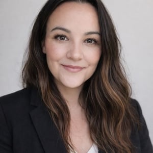 A professional headshot of a woman with long, wavy brown hair wearing a black blazer and smiling softly against a light background.