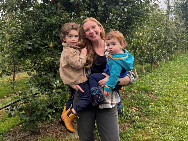 A woman in a plaid shirt holds two young children outdoors, surrounded by apple trees.