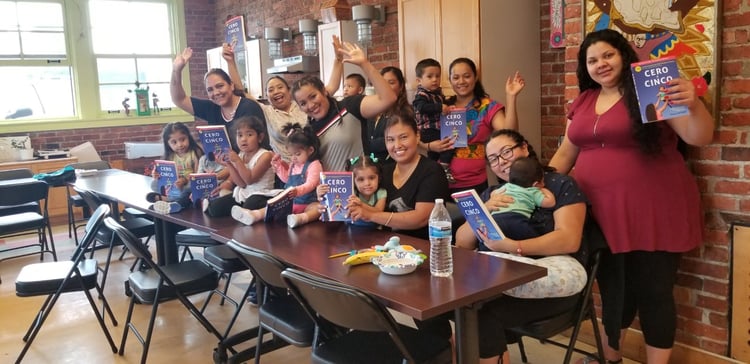 A group of several mothers and their children participating in the Spanish-language PEPS Group through Open Arms. The babies and toddlers are being held by their mothers or are sitting on a table. Many of them are holding a book called “Cero a Cinco.” 