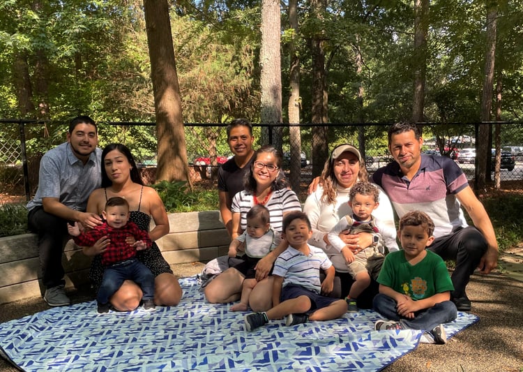 Three sets of parents sit outside with their young children on a picnic blanket. 