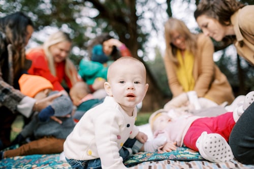A baby sitting on a blanket in a park looking at the camera with several babies and moms in the background. Photo credit: The Happy Film Company.&nbsp;&nbsp;