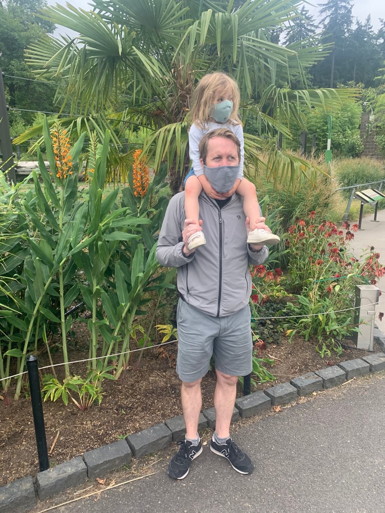 A toddler girl in a light blue face mask sitting up on her father’s shoulders as they both look to the left while standing in front of a lush green garden of palm trees and flowers.