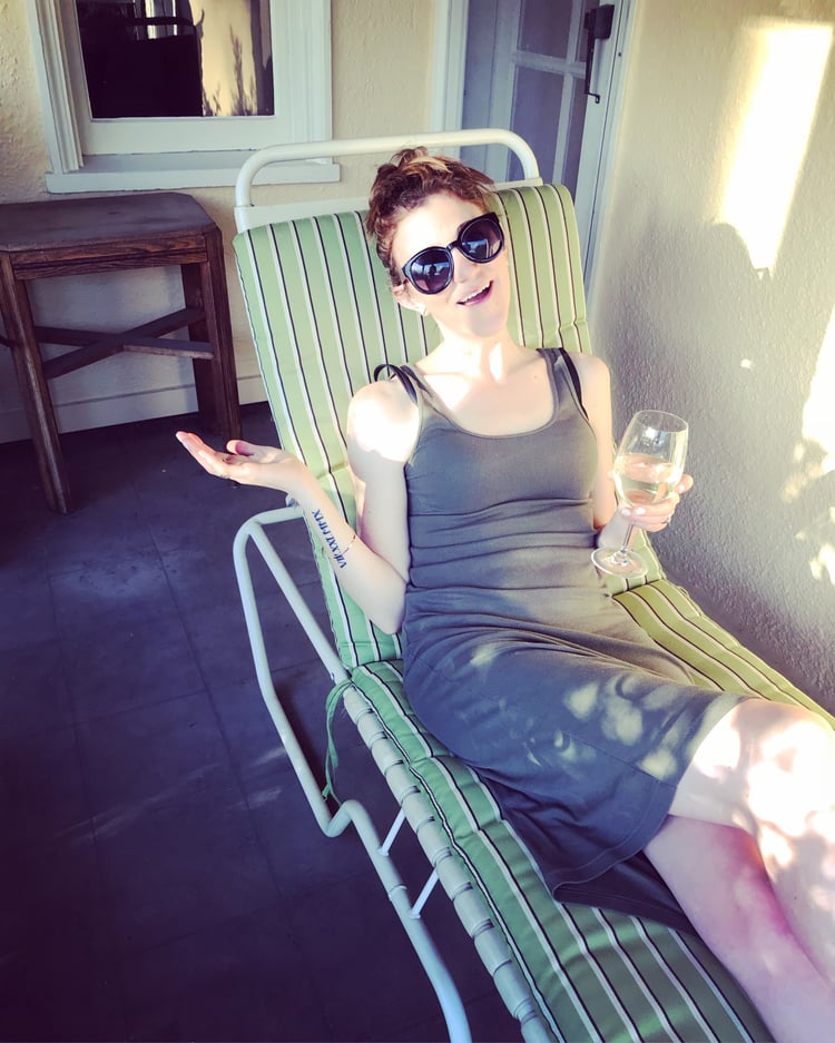 A woman wearing a pair of sunglasses, sitting on a patio chair, enjoying a glass of white wine in the sun. 