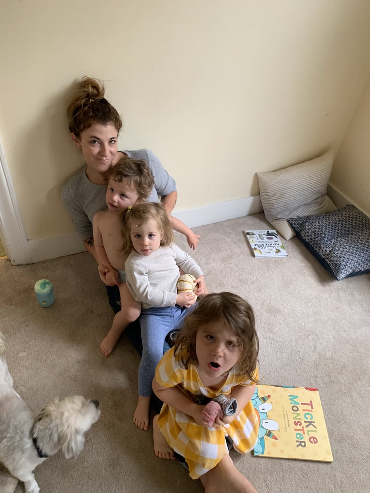 A mom with her three children sitting along her legs, reading a book. 