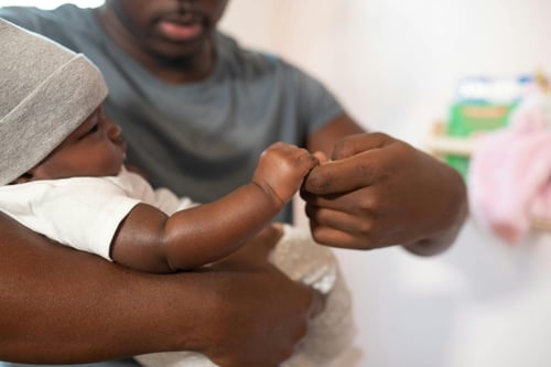A father holding his infant in his arms while the baby grasps the father’s finger. Photo credit: Nappy via Pexels.