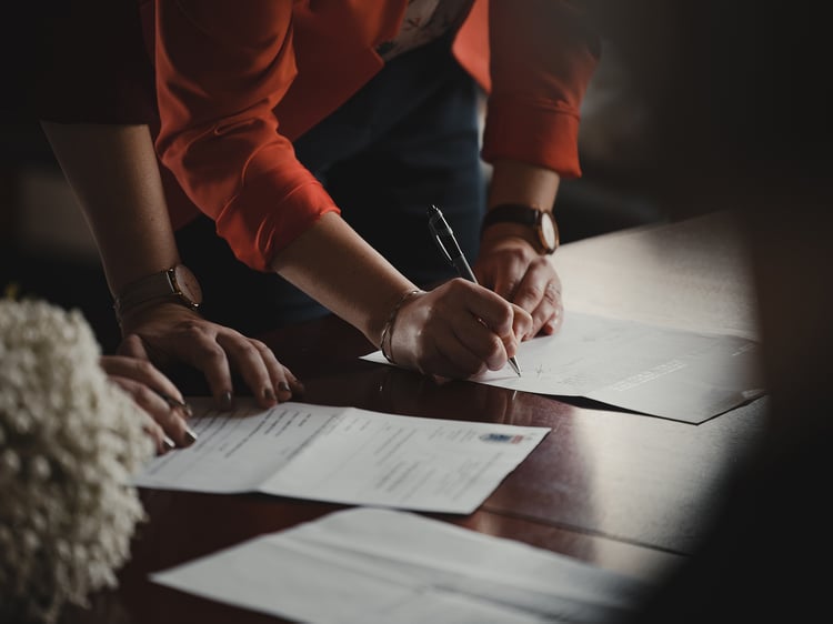 Two people standing over a table with white sheets of papers spread out on a dark wooden table and pens in hand, getting ready to sign documents.  